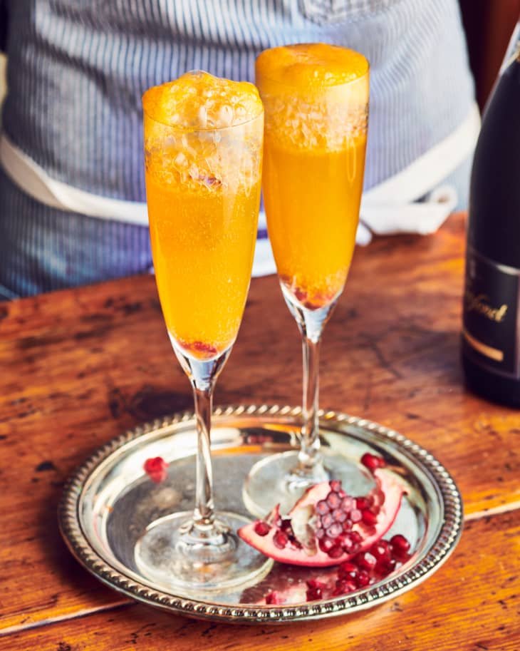 Two mimosas sit on a silver tray that is garnished with a pomegranate slice.