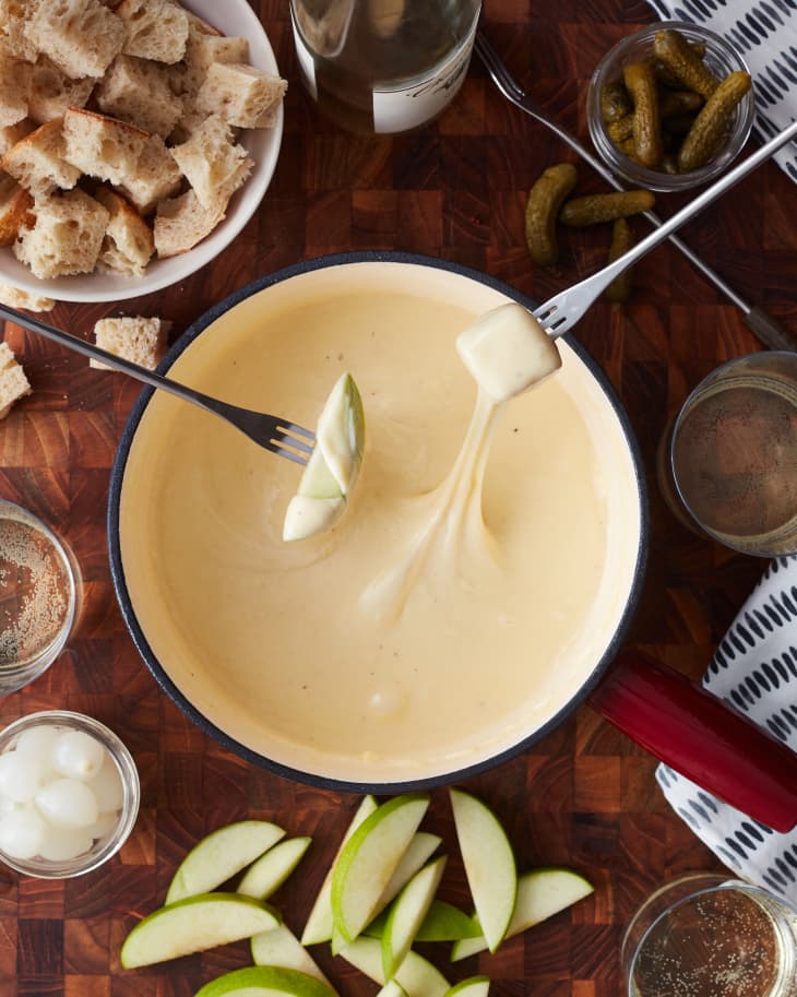 people are dipping a piece of bread and a sliced apple into the cheese fondue next to a bowl of bread and sliced apples