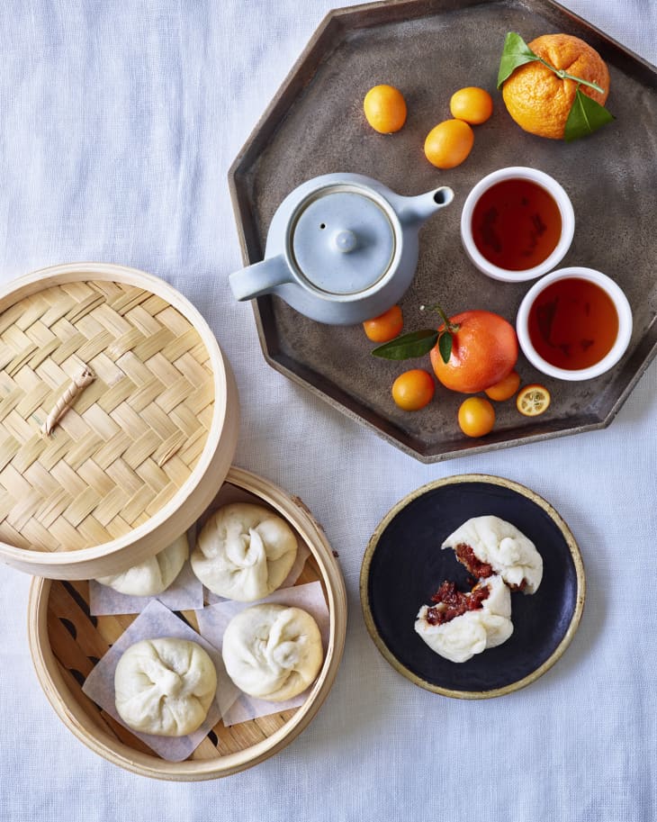 bao buns sit in the container, and on a plate next to a tea set with citrus