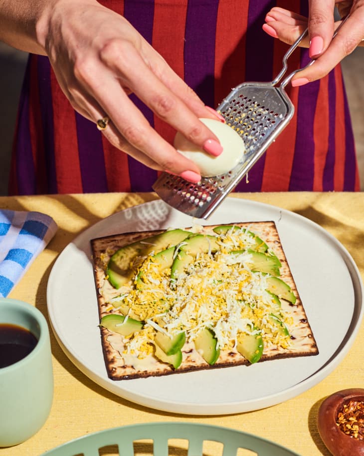 Matzo with Avocado &amp; Grated Egg, with egg being grated at the table, frontal view.