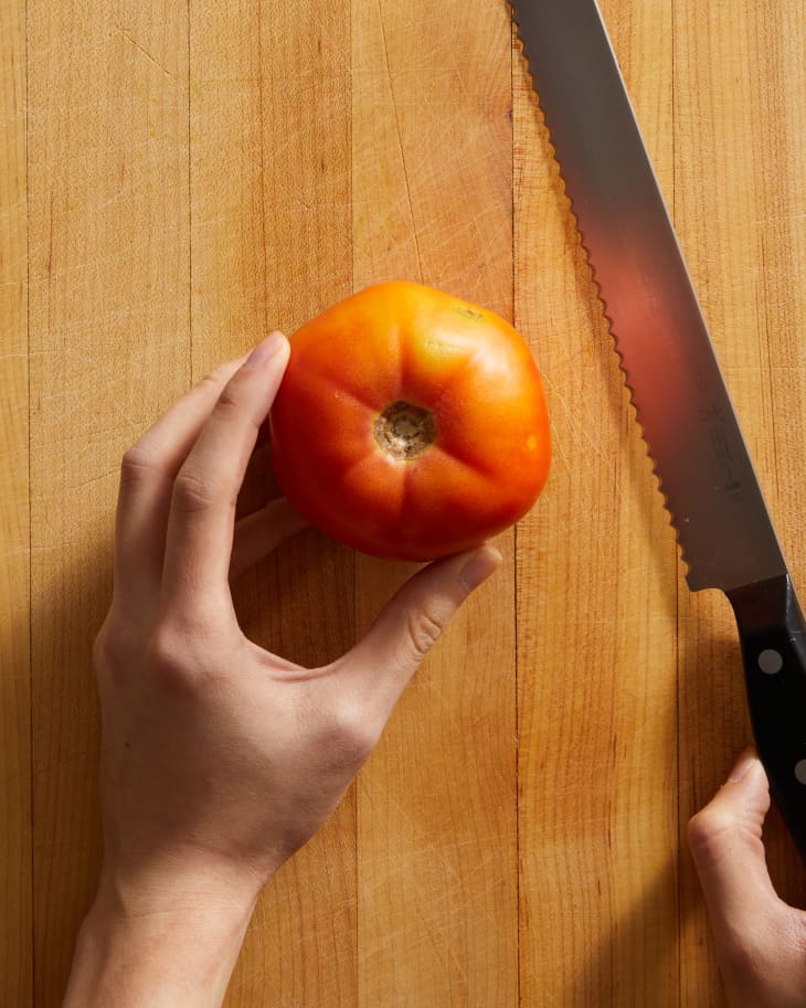 tomato on cutting board with someone holding a serrated knife