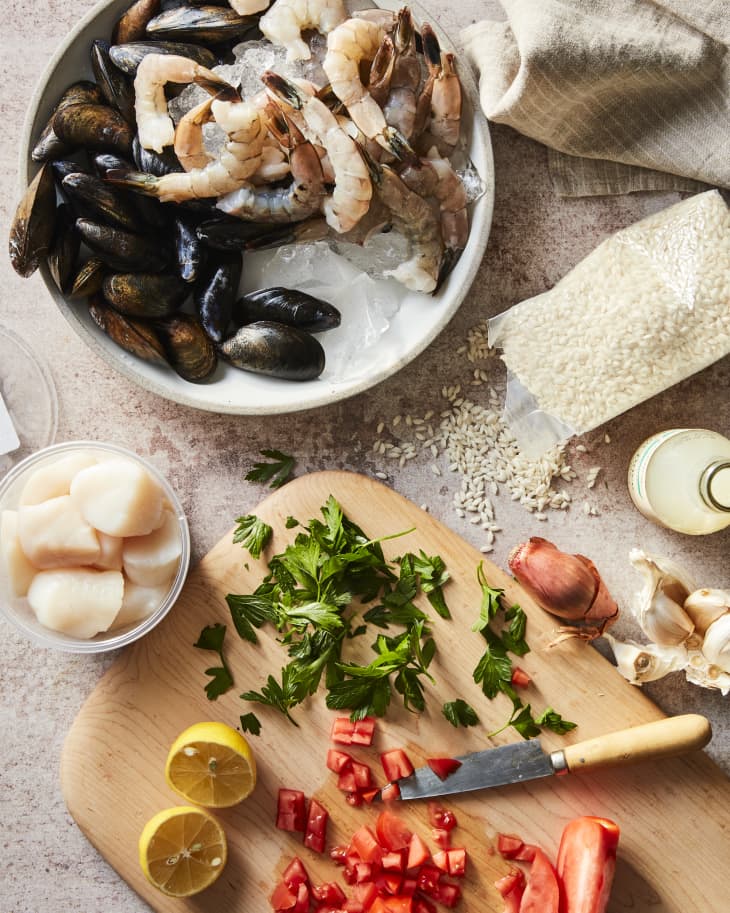 Prep scene for seafood risotto. A bowl of fresh mussels and shrimp on ice, a small glass bowl of scallops, a cutting board with chopped tomatoes, herbs, lemon. Some garlic and parmesan cheese on surface