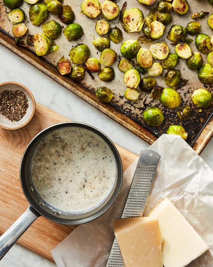 roasted brussels sprouts on sheet pan with saucepan of cacio e pepe sauce in frame. Cheese zester with parmesan cheese block in the lower right corner and ground black pepper in a small bowl. Prep moment