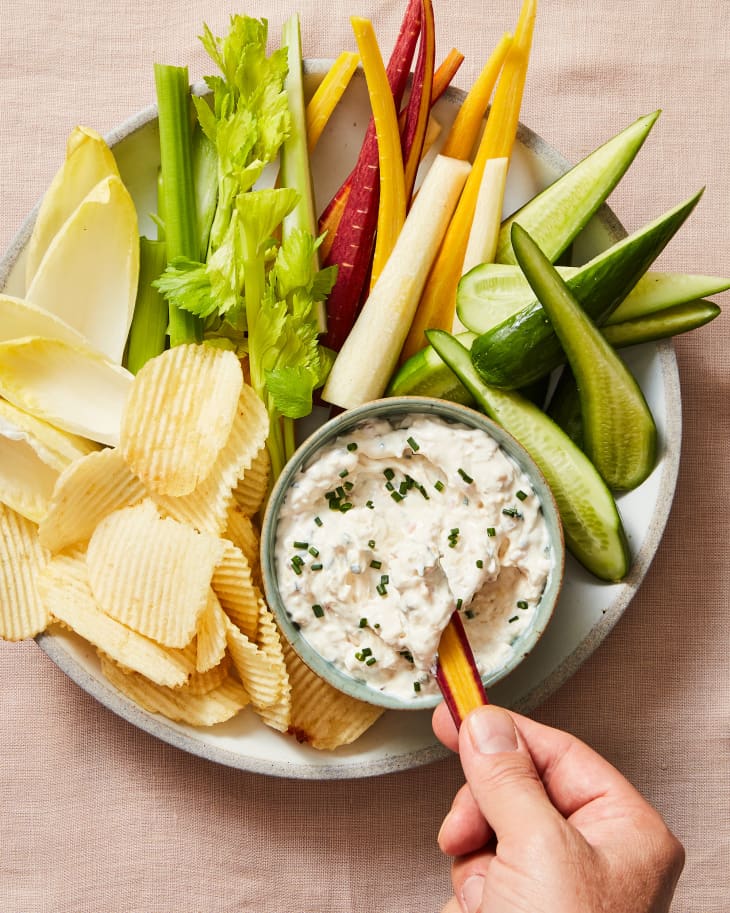 bowl of clam dip on platter with ridged potato chips and a variety of vegetables. Someone is dipping a carrot