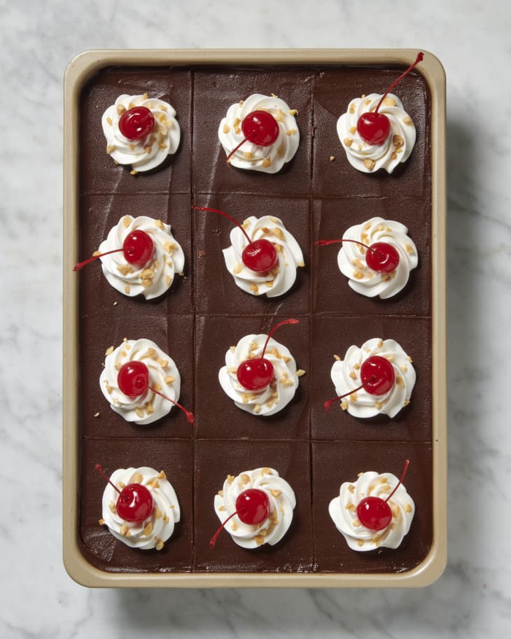 an overhead image of a whole banana split poke cake cut into pieces with chocolate frosting, whipped cream, nuts, and cherries on top