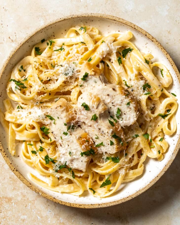 An overhead photo of a bowl of chicken fettucine Alfredo with grated parmesan cheese and parsley on top on a tan stone background.