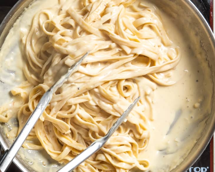 Pasta added to creamy alfredo sauce in a skillet, getting tossed with silver tongs.