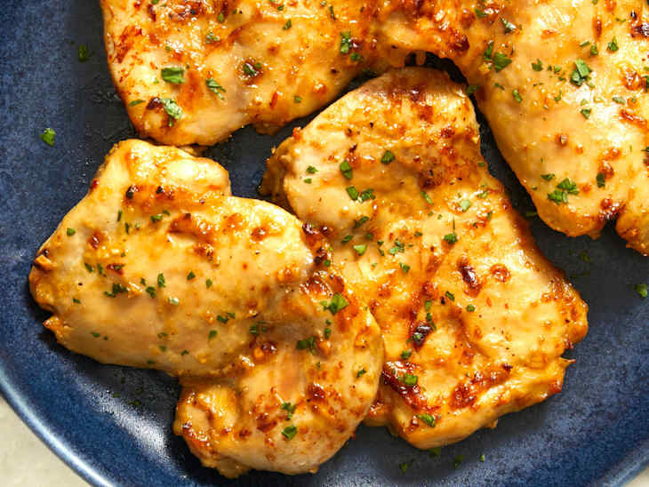 Close up of four pieces of baked boneless, skinless chicken thighs stacked on a dark blue plate on a marble background