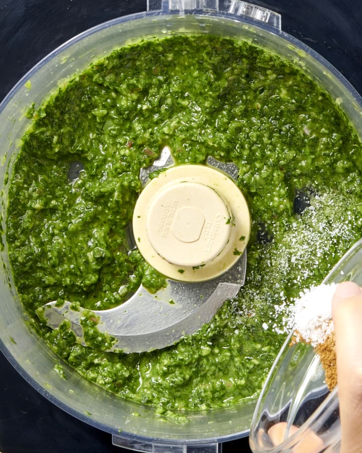 salt and seasoning being added to mixed herbs in a stand mixer in the process of making chimichurri
