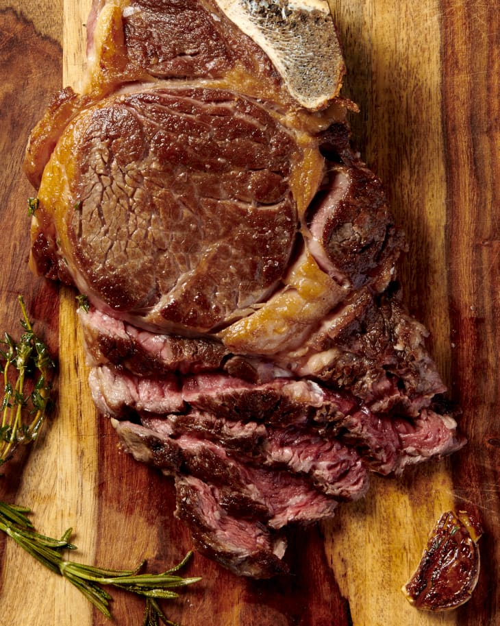 a medium-rare ribeye steak sliced on a cutting board with basting herbs and garlic