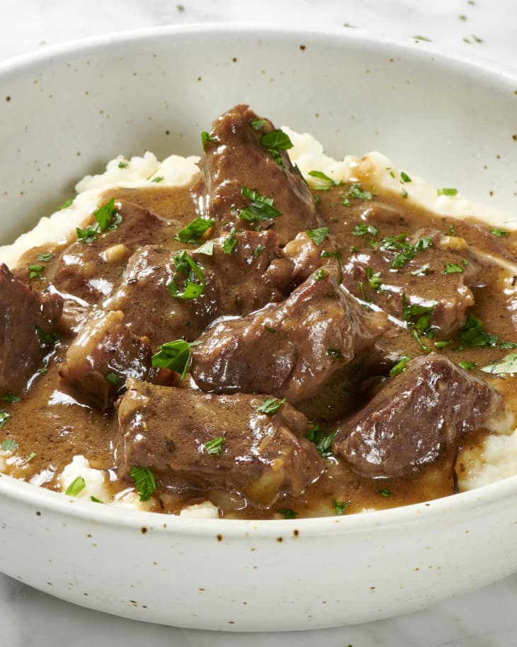 Angled view of beef tips on mashed potatoes, topped with herbs in a white and brown speckled bowl.