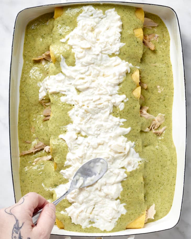 Overhead view of spoon spreading cheese mixture on top of enchiladas with green sauce.