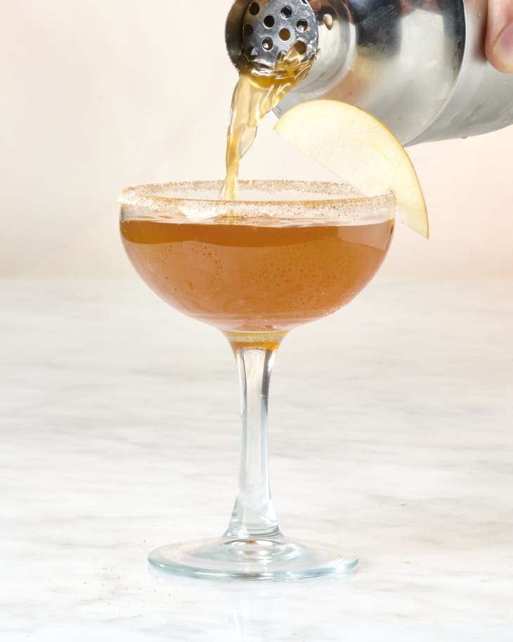 Harvest cocktail being poured into a single coupe glass with cinnamon sugar rim and apple garnish.