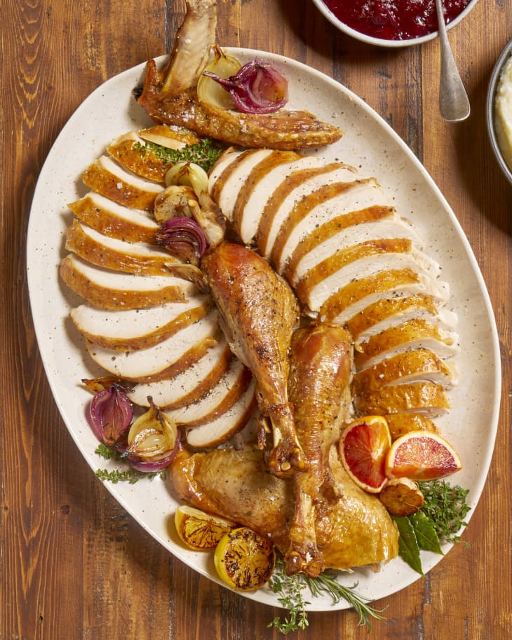 Overhead view of sliced turkey on a beige and brown ceramic platter with roasted blood orange slices, onions and herbs.