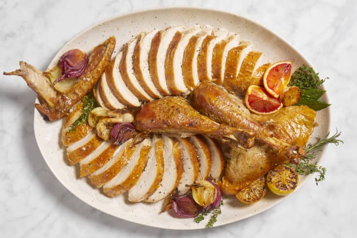 Overhead view of a sliced turkey on a beige and brown ceramic platter with roasted blood orange slices, onions and herbs.