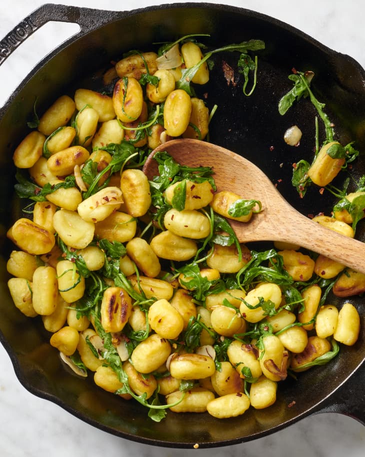 Overhead view of gnocchi and arugula in cast iron pan, being scooped with a wooden spoon.