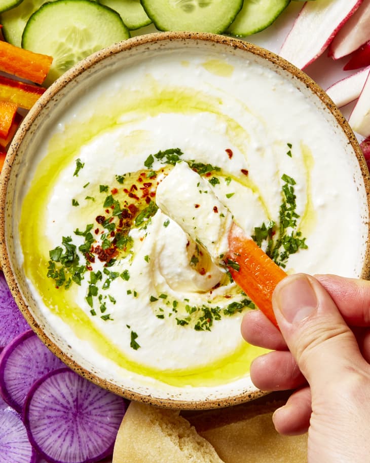 Overhead view of a small bowl of feta dip, topped with herbs and oil, on a plate surrounded but assorted cut vegetables and pita - and a hand dipping a carrot into it.