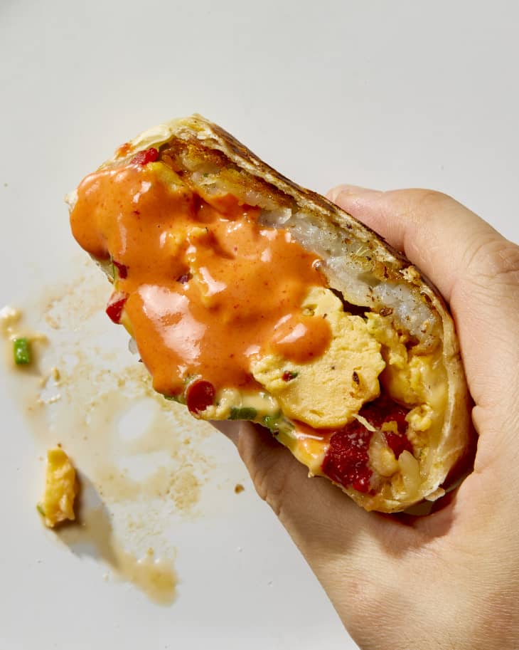 Overhead shot of a hand holding a half of the burrito with a lot of hot sauce on the corner of it.