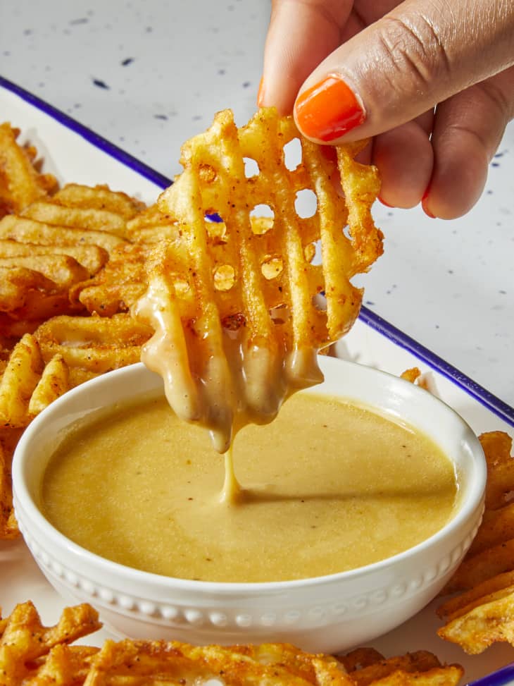 Head on shot of waffle fries, and a small white bowl of chick fil a sauce on a white dish with a blue rim. In the bottom top right of the image theres a hand dipping a waffle fry into the sauce.