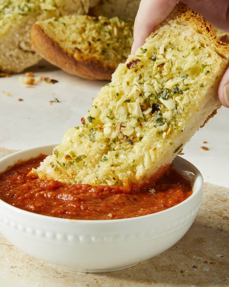 Head on shot of a hand dipping a slice of garlic bread into a small white bowl of marinara sauce.