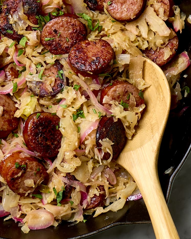 close up shot of cooked sauerkraut and sausage in a cast iron pan, with a wooden spoon resting in the bottom right part of the pan.