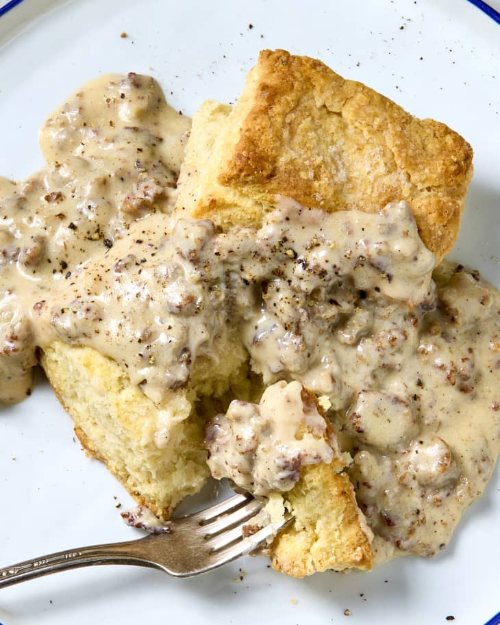 A plate with a split biscuit topped with sausage gravy and cracked black pepper with a bite on a fork ready to be eaten.