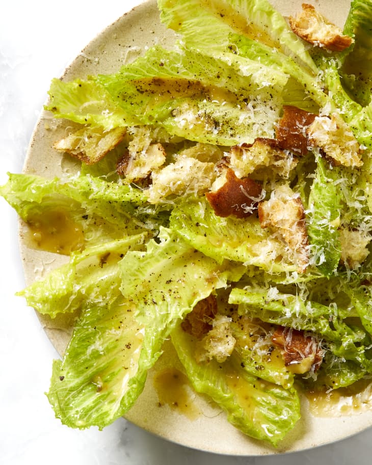 overhead shot of a caesar salad on a light beige plate, topped with croutons, grated cheese and black pepper.