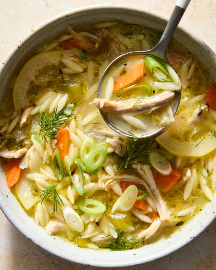 Overhead shot of chicken orzo soup in a white and brown speckled bowl, with a spoonful of soup hovering over the bowl.