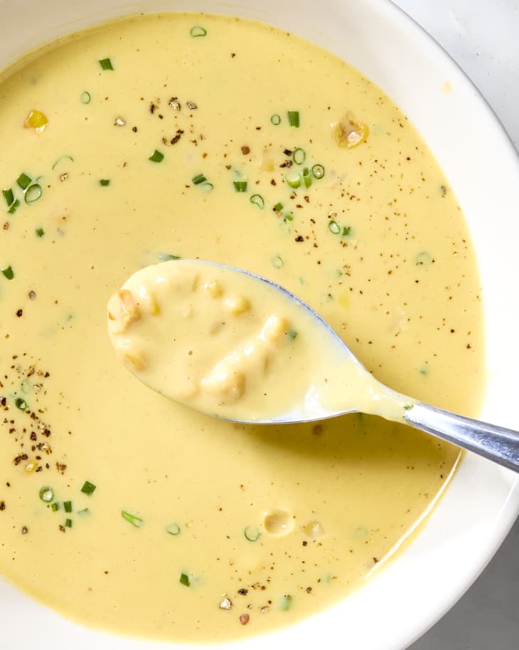 Overhead shot of corn soup in a white bowl, topped with green onion and black pepper. In the bottom right there is a spoon with a bite of soup resting in it.