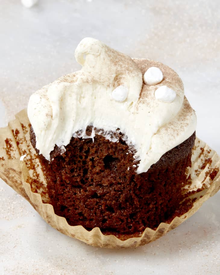 Close up shot of a single hot cocoa cupcake with a bite taken out of it and the wrapper coming off - dusted with hot cocoa powder and topped with mini marshmallows.
