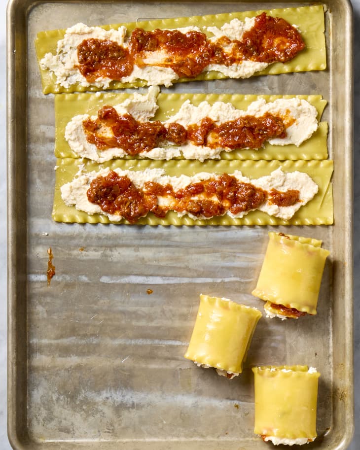 Sheets of lasagna pasta on a sheet pan topped with cheese and sauce with half rolled into a spiral.