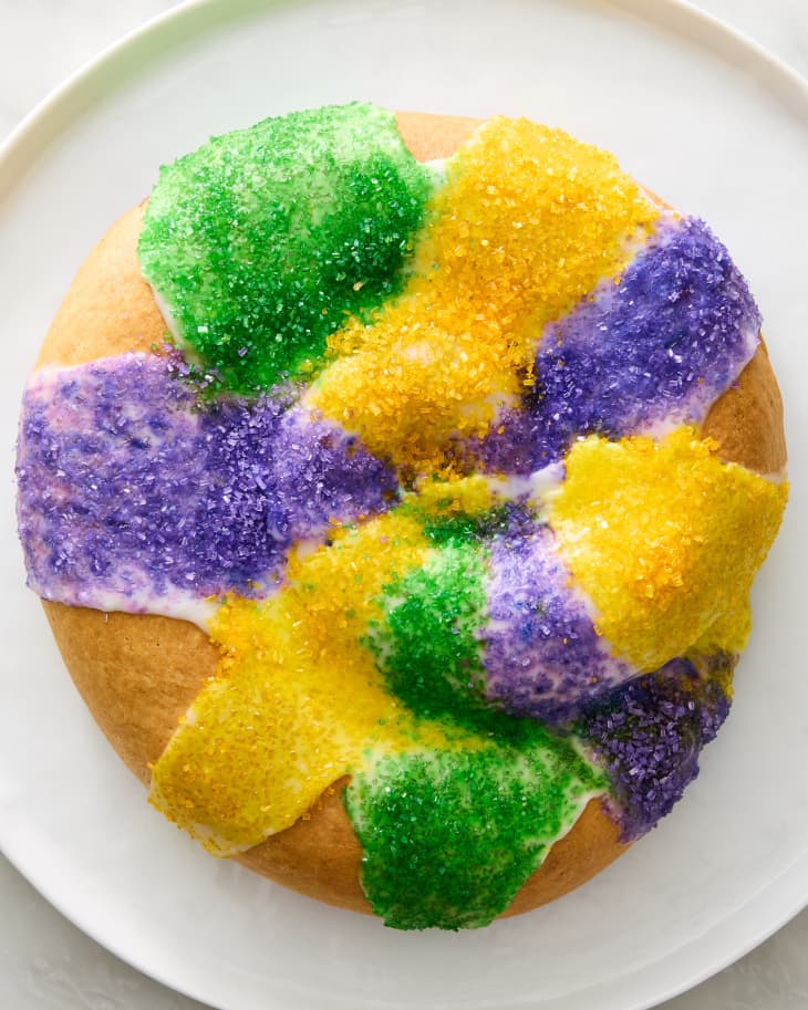 Overhead shot of a whole king cake on a round white platter. The cake is round with green, yellow and purple sprinkle stripes.