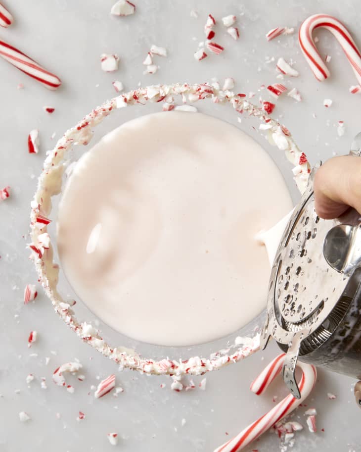 Overhead shot of someone pouring a peppermint martini into a martini glass with a candy cane rim.  On the surface below the glass is broken candy cane pieces scattered around.
