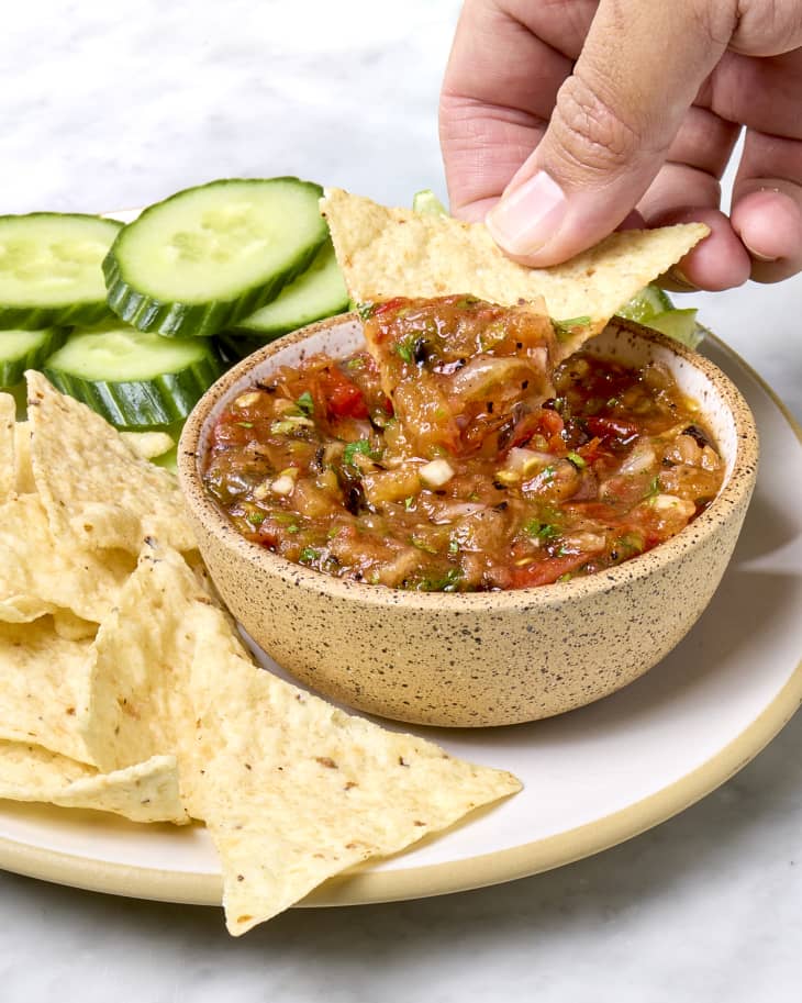 A bowl of ranchero sauce served with tortilla chips, sliced cucumber, and limes with a chip being dipped into the sauce.