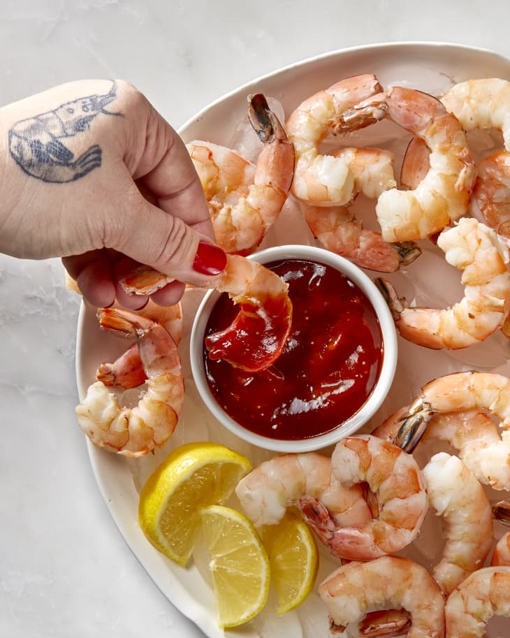 Overhead shot of a hand with a shrimp tattoo is dipping a cooked shrimp into the cocktail sauce that is in a small white bowl , resting in the larger bowl with the shrimp on ice.