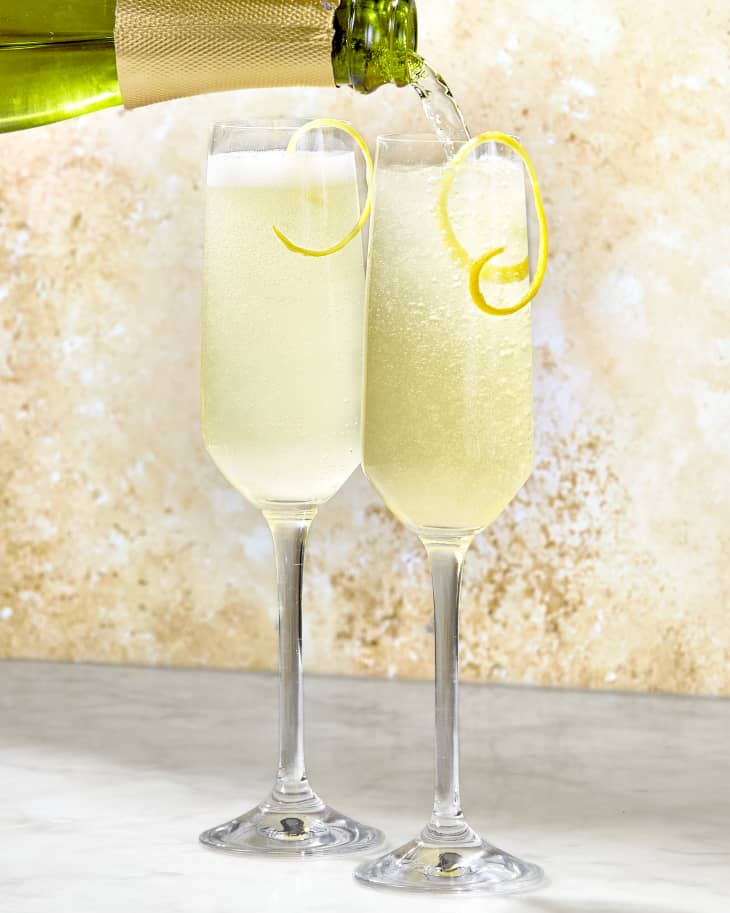 Two French 75 cocktails in champagne flutes on a marble surface against a warm stone background with one being topped off with a bottle of champagne.