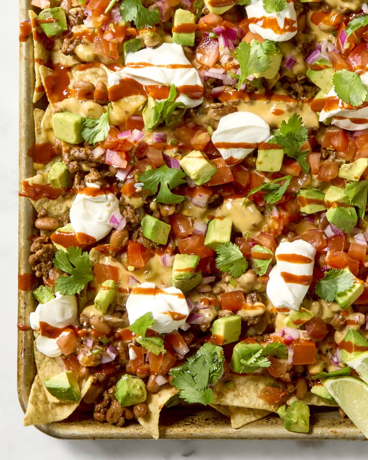overhead shot of loaded nachos on a baking sheet, topped with sour cream dollops, avocado and a drizzle of hot sauce.
