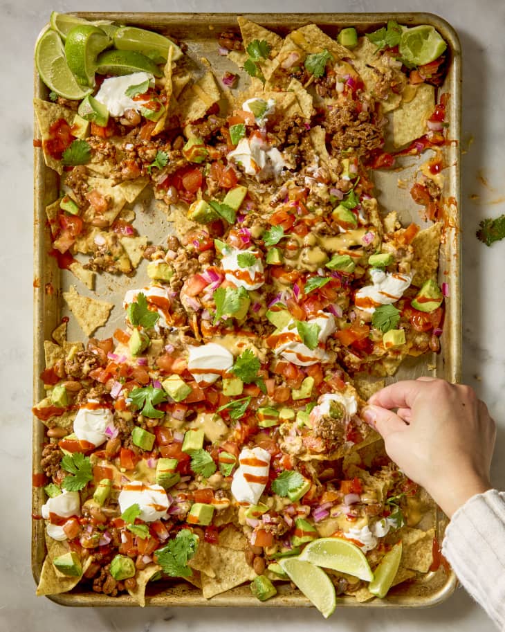 overhead shot of loaded nachos on a baking sheet, topped with sour cream dollops, avocado and a drizzle of hot sauce, with a hand grabbing a nacho off of the pan.