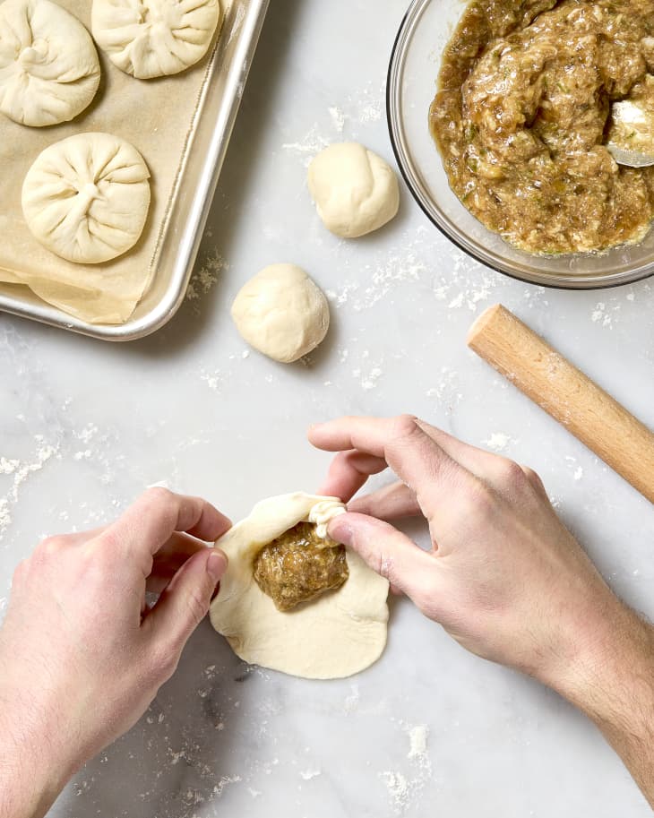 A photo of two hands folding dough around filling for pan fried pork buns.