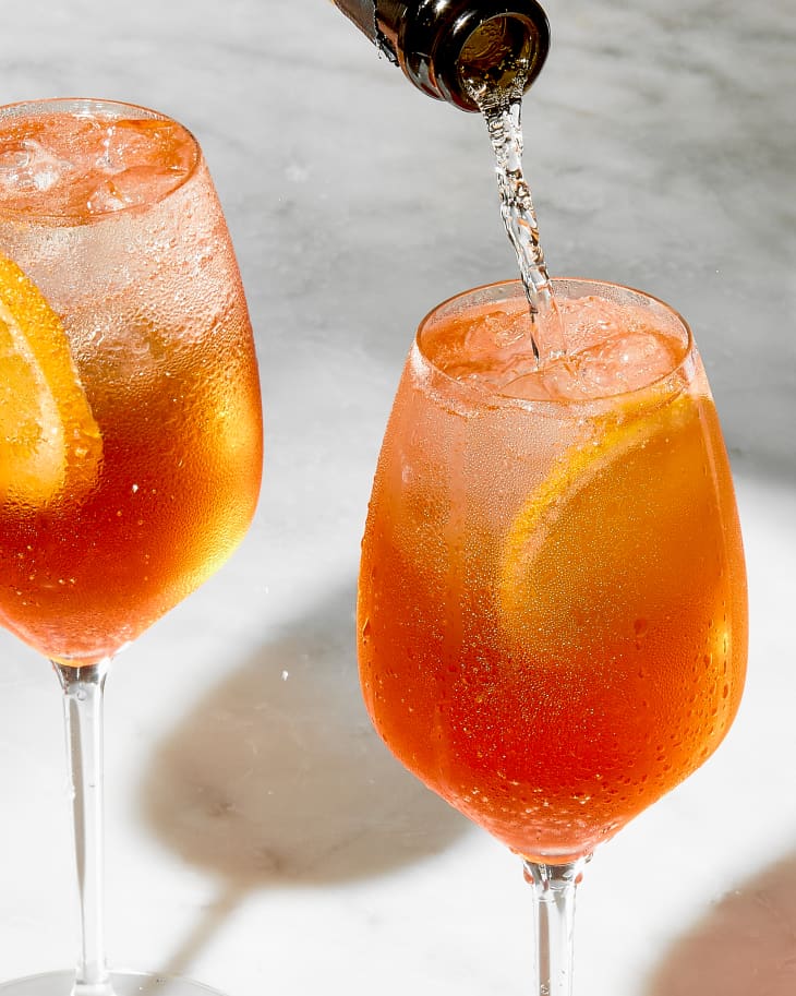 angled shot of two aperol spritz's in wine glasses, with prosecco being poured into one of the glasses.