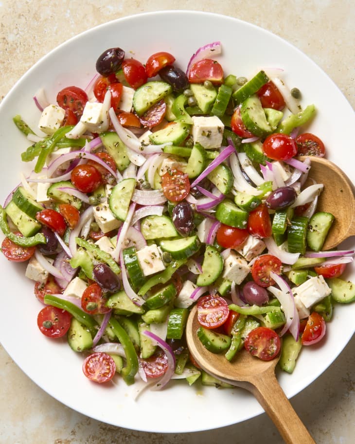 overhead shot of a greek salad in a large white serving bowl, with wooden serving utensils in the salad bowl