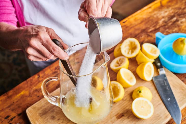 sugar being poured into a pitcher with lemon juice, stirring with wooden spoon