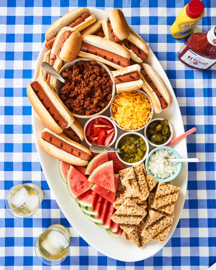 A tray of hot dogs sits on a picnic blanket. On the tray are hot dogs in hot buns, a bowl of ground beef, cheese, jalapeños, tomatoes, relish, watermelon, and grilled rice crisps.