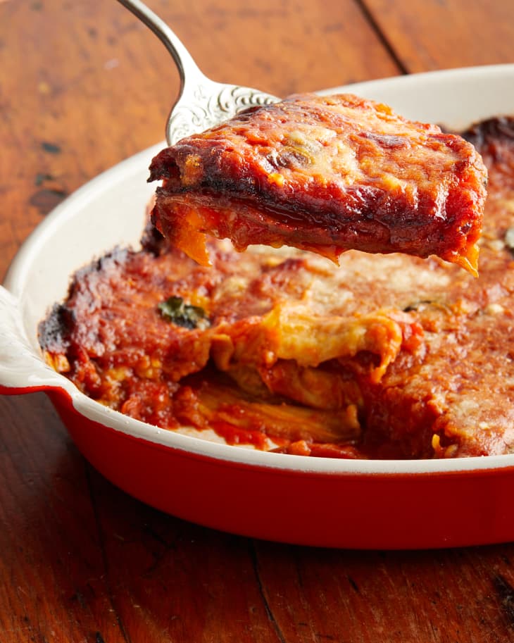 Eggplant parmesan baked in a shallow red dish with piece sliced out and placed on a serving knife.