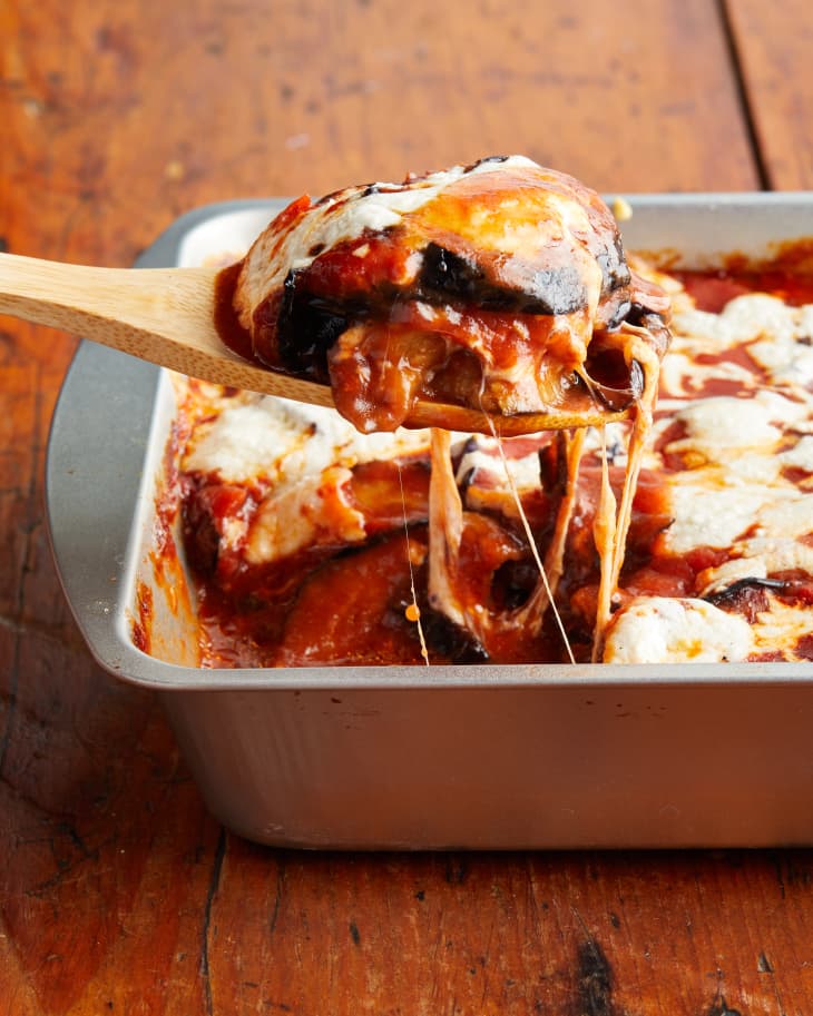 Baked eggplant parmesan in tin baking dish, piece being scooped out with wooden serving spoon.