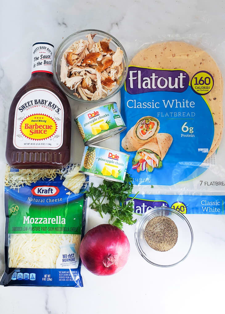 Flatbread ingredients laid out (clockwise): Flatbread packets, ground pepper in small glass bowl, red onion, mozzarella, barbecue sauce, shredded chicken, canned pineapple, fresh herbs.