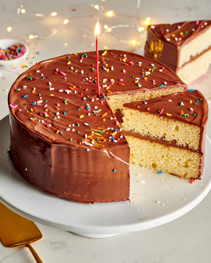 a slice of cake is missing from a circular birthday cake on a white tray with sprinkles and a candle on top