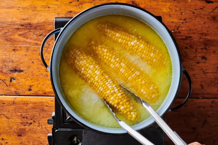 Tongs pull three ears of corn in a dutch oven filled with a  butter bath.