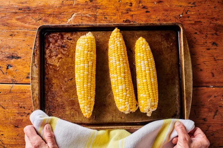 Three ears of corn are on a sheet pan and being placed on a wood table by two hands.