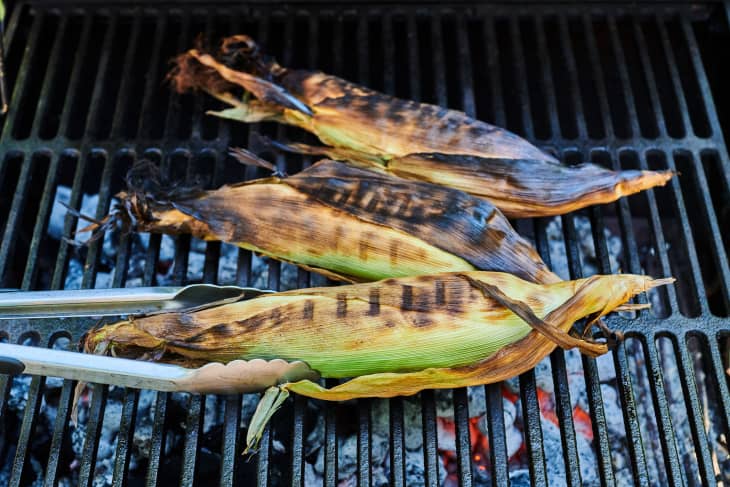Three ears of corn, still in the husk, are grilled on an outdoor grill.